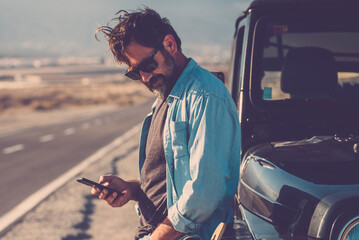 Traveler man use phone mobile cnnection standing against a black off road car with long road in background. Road trip and travel lifestyle for adventurer male people. Use of digital map road trip - obrazy, fototapety, plakaty