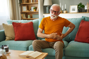 Obraz premium Senior man suffering from stomach ache sitting on a couch in the living room at home