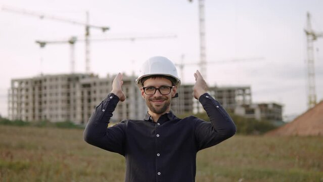 Caucasian happy construction engineer wearing glasses masterfully and deftly puts on helmet. Male manager is ready to work and start sales. Safety precautions on construction site.