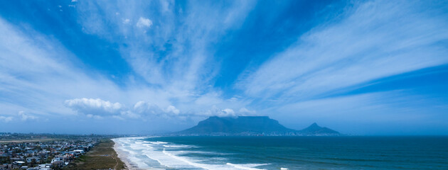 Aerial wide view of Table Mountain in Cape Town on a sunny day, clouds billowing over the mountain and beach in foregound.
