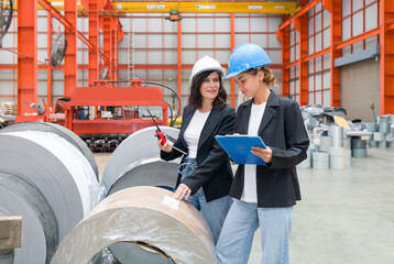 Two ladies with safety helmet are observing metal roll inside an industrial site.