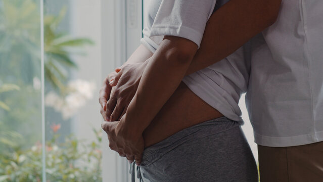 Young Asian Pregnant Couple Hug And Holding Belly Talking With Their Child. Mom And Dad Feeling Happy Smiling Peaceful While Take Care Baby, Pregnancy Near Window In Living Room At Home Concept.