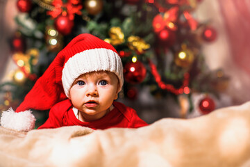 Joyful baby looking at camera in Christmas photo. Newborn baby in red cap of Santa Claus and Christmas tree in background