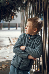blond boy near a wooden fence in the autumn city. dressed in a blue sweater and jeans smiling and looking away