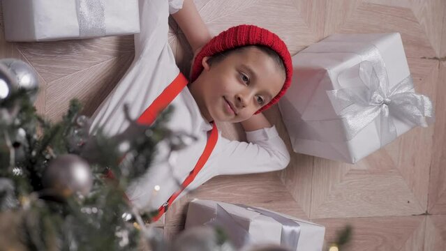 Merry Christmas kid. Portrait of a happy funny cute baby boy child one 6 years old in red clothes a boy lying on the floor under the tree amongst boxes of presents. New Year's Eve.
