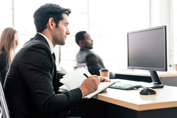 The Caucasian businessman jot something on small notebook while looking on computer monitor at his desk workplace. Positive and optimistic mood of businessman. Photo with copy space.