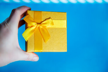 Hand holding a gold gift box with a yellow ribbon on blue background copy space close-up