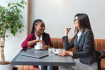 Two young business woman colleagues taking a break in nearby cafeteria drinking coffee talking about private life, to get know each other better. Staff members company employees sitting in the cafe