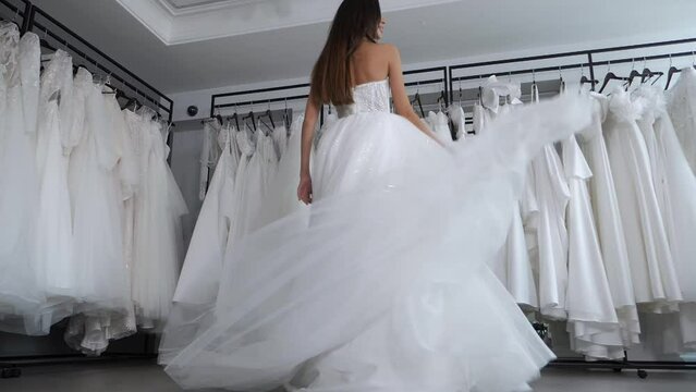 A gorgeous slender bride with long hair is spinning in a wedding dress in a wedding salon, a woman is trying on. Slow motion. Stabilized camera image from below. Wedding salon with lots of dresses.