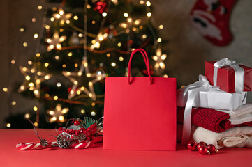 Red shiny shopping back mockup on table, red and white gift boxes, sweaters, candy canes on red table, blurred Christmas tree background. Holiday presents, winter shopping sales, branding template