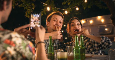 Friends Enjoying a Summer Evening at a Caravan Camping Area. Fooling Around, Joking, Eating Pizza. Lovely Couple Posing for a Funny Photo with Them Biting a Slice of Pizza - obrazy, fototapety, plakaty
