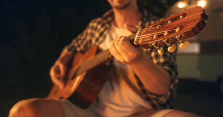 Close Up Young Man Spending Time at a Campfire, Sitting in Chair and Playing Guitar Music at a...