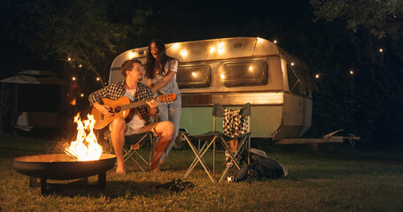 Portrait of Friends Having Fun, Playing Guitar Music and Dancing Outdoors at a Motorhome Rest Area. Loving Couple Enjoying a Summer Evening while Travelling and Camping in a Caravan - obrazy, fototapety, plakaty