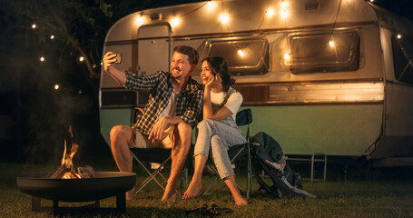 Loving Partners Using a Smartphone to Take Selfie Photos of Them Enjoying a Summer Evening at a Caravan Camping Area with Campfire. Young Couple Travel Together and Live in a Motorhome - obrazy, fototapety, plakaty