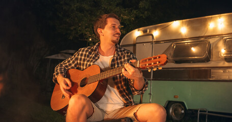 Portrait of a Young Man Spending Time at a Campfire, Sitting in Chair and Playing Guitar Music at a...