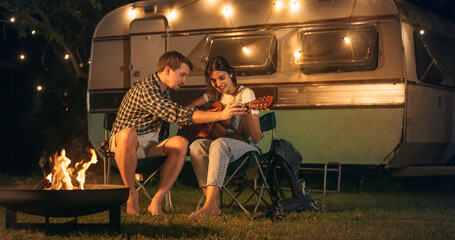 Couple Relaxing Next to Campfire in the Evening, Young Female Learning to Play Music on Acoustic Guitar. Partners Travelling Together, Living Outdoors in a Camper Motorhome