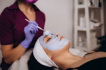 Cosmetologist applying blue mask to face woman client, rejuvenation procedure to beautiful in a...
