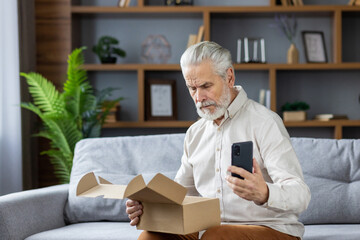 Shocked senior gray-haired man sits on the sofa at home, holds the phone in his hands and looks...