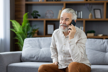A gray-haired senior man is sitting on the sofa at home and talking on the mobile phone