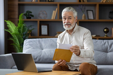 Fototapeta premium Portrait of an older gray-haired man sitting at home on the couch, holding a envelope with a letter he received in the mail, and looking at the camera