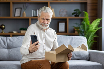 Senior man sitting at home on sofa and holding online purchase in box. Compares the product with the order on the phone, looks disappointed and upset at the screen - Powered by Adobe