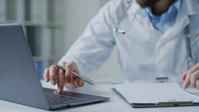 Closeup pan shot of unrecognizable male doctor looking at information in laptop and writing it down in medical history during workday in hospital