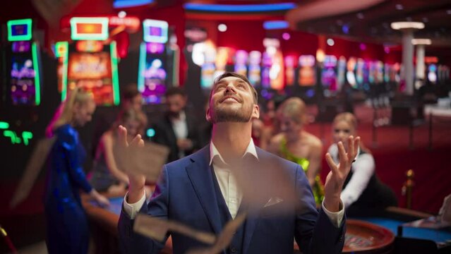 Happy Man in a Suit Standing in the Middle of a Modern Casino, Extending Arms, Welcoming a Money Rain.Young Male Smiling, Celebrating His Victory. Embodiment of a Successful Man
