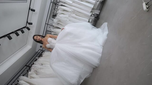A happy slender woman poses in a wedding dress in a wedding salon against the background of many different dresses, she holds a long skirt and rotates, the camera rotates 360 degrees. 3D roll.