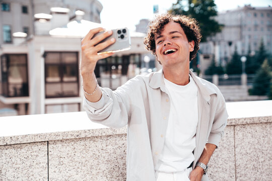 Portrait of young attractive man with curly hair hairstyle. Smiling handsome male in casual stylish clothes posing in the street at sunny day. Cheerful and happy model outdoors. Take selfie photos