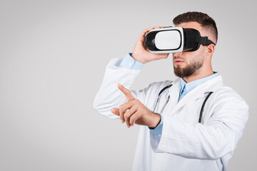 Young male doctor in white coat using VR glasses, grey background