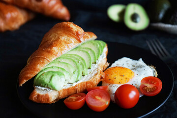 Croissant with avocado, scrambled eggs and cherry tomatoes on a black plate next to vegetables and a fork on a dark background