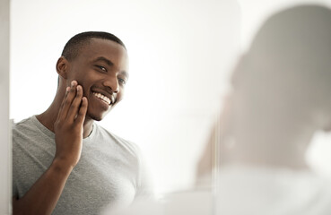 Smiling African man examining his face in a bathroom mirror - obrazy, fototapety, plakaty