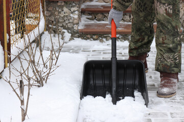 A man in gray gloves removes snow with a shovel with an orange handle from a path made of gray...