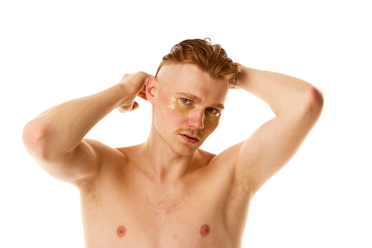 Young man styles, combing hair looking at camera with hydrogel patches under eyes against white studio background.