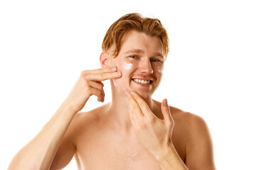 Anti-aging and skin-care products. Portrait of redhaired man applies face cream looking at camera with against white background.