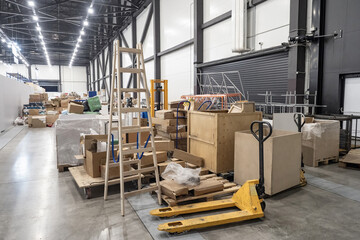Hangar with boxes and stepladder. Industrial premises with pallet jack near containers. Factory hangar with boxes filled with building materials. Industrial warehouse. Mess in storage space - obrazy, fototapety, plakaty
