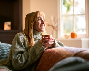 Smiling Woman At Home Wearing Winter Jumper With Warming Hot Drink Of Tea Or Coffee In Cup Or Mug