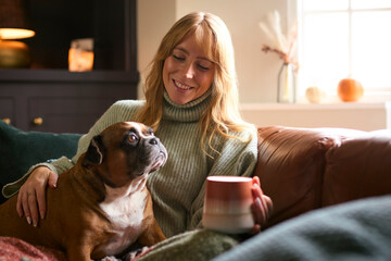 Woman At Home In Winter Jumper With Hot Drink Of Tea Or Coffee In Cup Stroking Pet French Bulldog