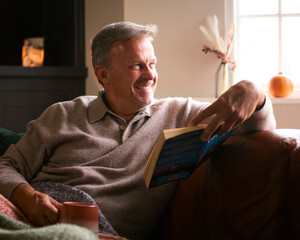 Mature Man At Home In Winter Jumper On Sofa With Warming Hot Drink Of Coffee In Mug Reading Book