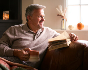Mature Man At Home In Winter Jumper On Sofa With Warming Hot Drink Of Coffee In Mug Reading Book