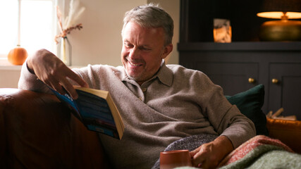 Mature Man At Home In Winter Jumper And Blanket With Warming Hot Drink In Mug Reading Book