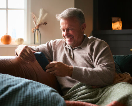 Mature Man At Home Sitting On Sofa With Warm Blanket Streaming To Mobile Phone