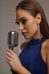 Stylish and beautiful female artist in a blue dress sings into a microphone on a gray background