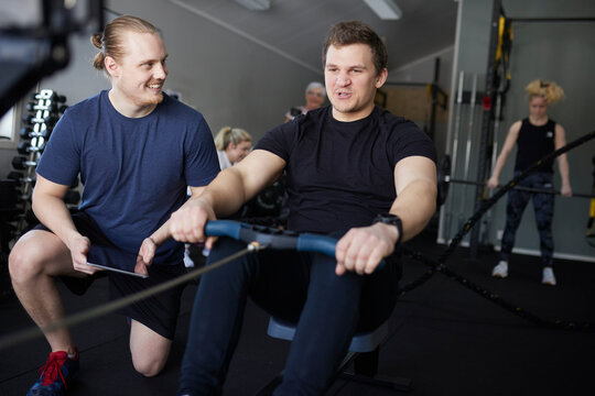 Smiling male instructor encouraging man exercising on rowing machine at gym