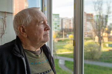 Portrait of a sad elderly man looking out the window at home. Lonely elderly man looking out the window