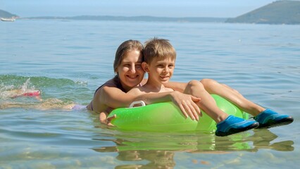 Fototapeta na wymiar Happy cheerful boy and mother swimming with inflatable ring in calm sea waves towards the beach