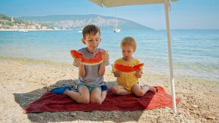 Two adorable boys enjoy sweet watermelon on the sea beach. Essence of a summertime getaway, holiday merriment, and the simple pleasures of vacation