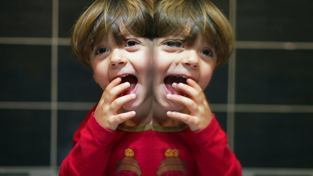 Humorous child creates twin image in mirror, joyfully peers at camera next to his mirrored self with happy expression