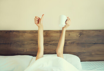Cheerful lazy woman waking up after sleeping lying in soft comfortable bed showing empty cup coffee stretching her hands up from under the blanket in bedroom at home