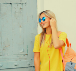 Stylish modern blonde young woman listening to music in headphones looking away wearing sunglasses, backpack, yellow t-shirt in the city